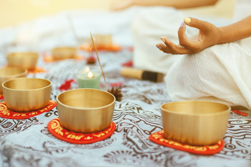 Misy tybetańskie ustawione w rzędzie na dekoracyjnym materiale, obok zapalonej świecy i kadzidła; w tle dłoń osoby medytującej w geście mudry, całość w ciepłym, złotym świetle tworzącym spokojną, duchową atmosferę.