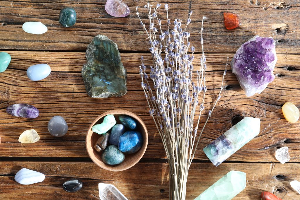 Naturalne kryształy i kamienie szlachetne ułożone na drewnianym tle, obok suszonej lawendy; widoczne m.in. ametyst, labradoryt, fluoryt i wygładzone minerały w odcieniach fioletu, zieleni i błękitu.