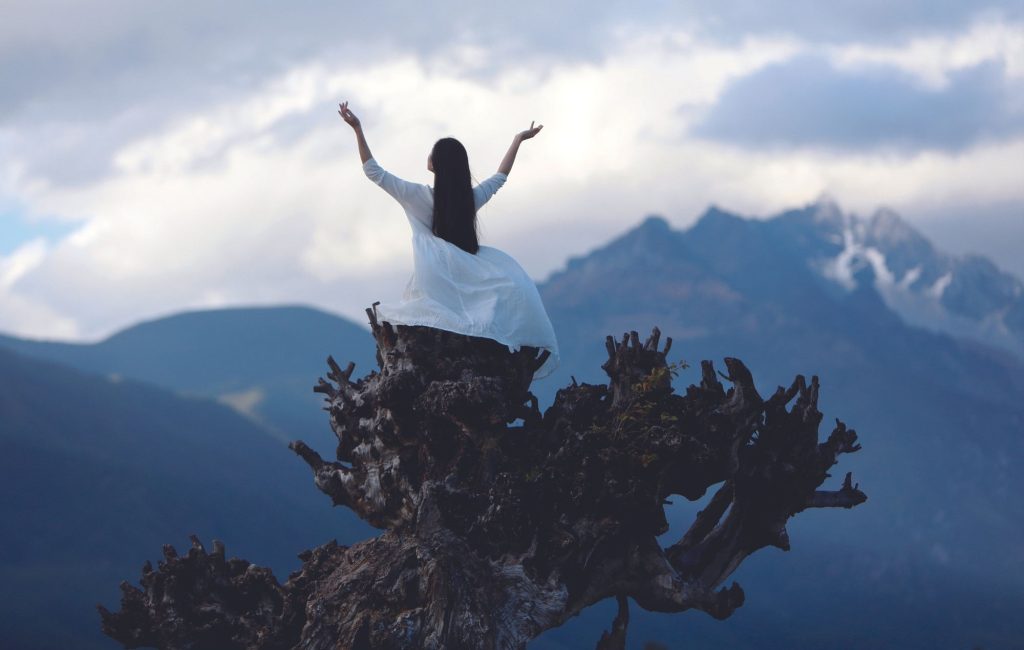 Kobieta w białej, zwiewnej siedzi na pniu drzewa na tle gór i chmur. Ma uniesione ramiona w geście wdzięczności.
