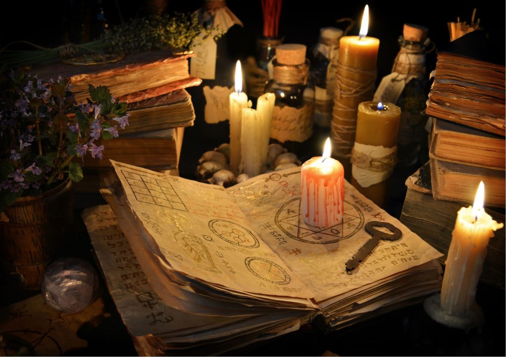 Stara księga zawierająca magiczne symbole i ich znaczenie. Na księdze stoi zapalona, czerwona świeca. Wokół znajdują się książki oraz świece.